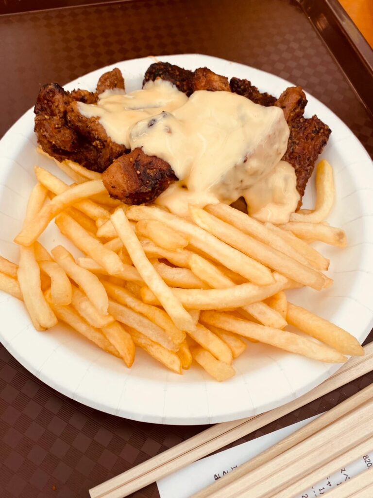 チーズスペアリブ Mサイズ。黒胡椒の効いたスペアリブにとろけるチーズをかけ、フライドポテトを添えた紙皿。トレイの上に割り箸が置かれている